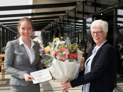 Bloemen voor de 100000ste bezoeker van het Afsluitdijk Wadden Center. Er zijn twee vrouwen in beeld. Zij staan in het restaurant van het museum. De ene vrouw houdt een bos bloemen vast en de andere een briefje met onleesbare tekst.