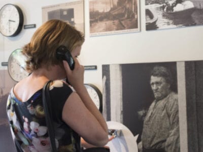 Expositie in het Zuiderzeemuseum over de Afsluitdijk. Een mevrouw staat in de expositiehal. Zij kijkt naar de wand, daar hangen oude foto's. In haar hand houdt zij een telefoon waaruit informatie klinkt.