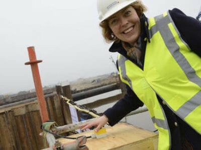 Gedeputeerde Sietske Poepjes op de Afsluitdijk. Zij staat scheef in beeld met een veiligheidshesje en helm. Zij heeft haar hand bij een knop waarmee een apparaat in werking wordt gesteld.