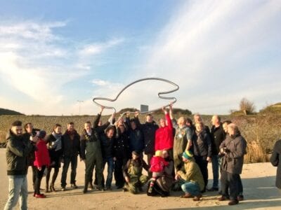 Groepsfoto bij het beeldmerk van de Blije Vis. Op de achtergrond zie je een soort duinen.