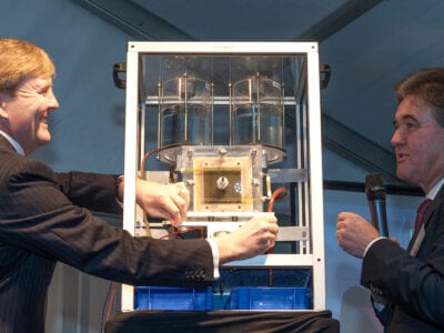 Koning Willem Alexander bezoekt Blue Energy. Hij staat aan een kleine versie van de Blue Energy technologie. Er staat nog een man omheen.