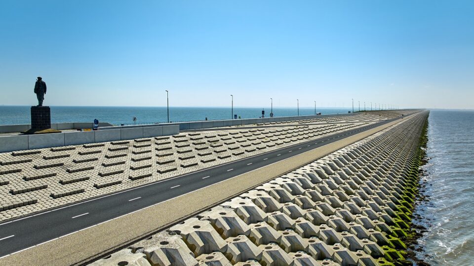 Schuin zicht op de Afsluitdijk, van linksonder naar rechts midden in beeld, onder een strakblauwe lucht. Onder en boven aan de dijk zijn verschillende betonblokken te zien. Tussen beide delen ligt een wandel- en fietspad. Links staat het standbeeld van ir. Lely.