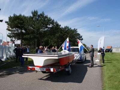 Pitchstop Dutch Solar Challenge. Je ziet een weg met mensen eromheen. Op de weg staat een boot en twee mensen houden een Nederlandse en Friese vlag vast.