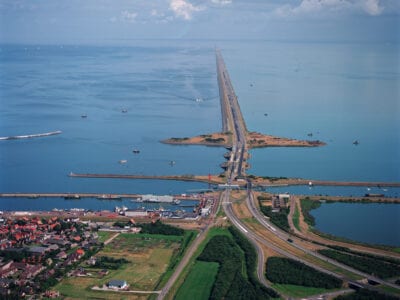 Luchtfoto van Den Oever. Wij kijken uit over de weg van de Afsluitdijk. Onder in de foto zien we een deel van Den Oever en de haven. De weg loopt dan verticaal uit beeld en is omringt met water.