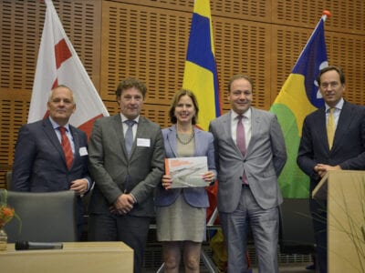 Bijeenkomst met genodigden voor het bidbook van de vebreding van de sluis bij Kornwerderzand. Wij zien op de foto vijf hooggeplaatste mensen staan. Een mevrouw in het midden heeft een boek vast. Op de achtergrond zien we drie kleurige vlaggen.