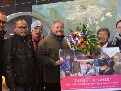 Foto van de 10.000ste bezoeker voor het oude informatiecentrum op de Afsluitdijk. Er staat een rij met zeven mensen en zij hebben een cheque vast.