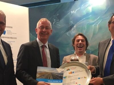 Overhandiging van het Bidbook: De Nieuwe Afsluitdijk. Er staan vier mannen naast elkaar die een schotel en boek omhoog houden.