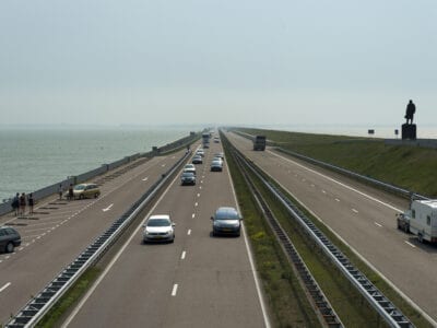 Verkeer op de Afsluitdijk tussen Noord-Holland en Fryslân. Wij kijken van boven op de twee weghelften. Aan de rechterkant staat het standbeeld van Lely.
