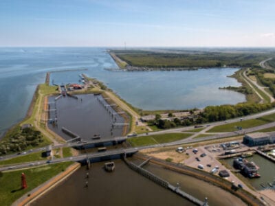 Luchtfoto van de dijkring bij Wieringen. We kijken uit op de dijkring, dit is een landstrook die verticaal op de weg van de Afsluitdijk staat. Aan de rechterkant zien we een kleine haven.