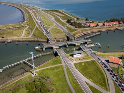 Luchtfoto van de spuihaven. We kijken van boven op de weg bij de Afsluitdijk. Er lopen twee weghelfte verticaal uit beeld. Om de weg zien we gras en een paar huizen staan. De brug staat open en er vaart een boot doorheen.