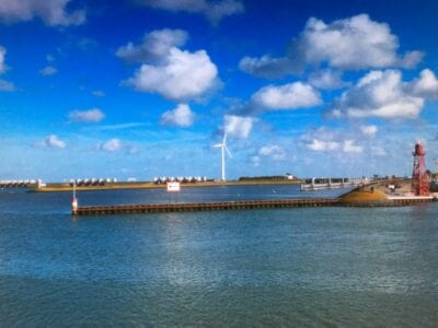 Vuurtoren bij Den Oever. Wij zien een landtong die in het water ligt. Op de achtergrond staat een witte windmolen. Op de landstrook staat een rode vuurtoren.