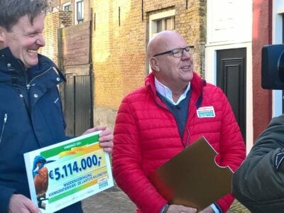 Uitreiking van cheque voor laatste kilometer van de Vismigratierivier. De cheque bedraagt 5 miljoen euro. Gaston staat in een straat met een man met een cheque. Er staat een man met camera voor hem.