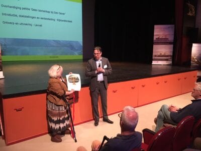 Tijdens de presentatie van Rijkswaterstaat over het ontwerp van de Afsluitdijk, werd een petitie tegen bomenkap aangeboden. Dit werd aangeboden door een mevrouw aan een meneer met microfoon. Zij staan voor een podium.
