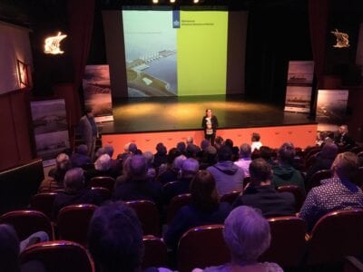 Presentatie van Rijkswaterstaat over het ontwerp van de Afsluitdijk, met veel publiek. Wij kijken van boven, over de hoofden van het publiek. Voor een podium staat een mevrouw. Achter haar een scherm met een presentatie van Rijkswaterstaat.