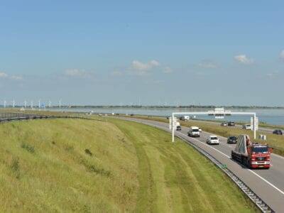 Verkeer op de Afsluitdijk. Wij kijken uit over het gras van de Afsluitdijk. Rechts zien we de weg met verkeer, nog verder het water.