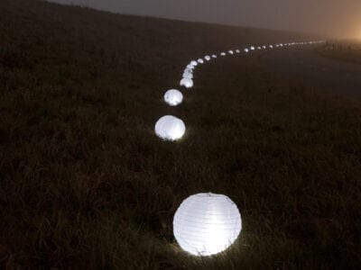 Duizenden lampionnen verlichten de Afsluitdijk. We zien de lampioenen in het gras van de dijk liggen. De rest is donker.