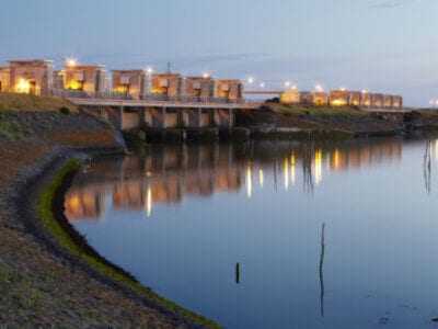 De Lorentzsluis. We kijken uit over het water, aan de linkerkant zien we de Afsluitdijk omringt met de sluisgebouwen. Deze zijn verlicht want de schemering valt in.