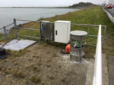 Opstelling van de tachymeter met beschermkooi. Wij zien een kooi met daarin de opstelling. Dit zijn een paar kastjes en een ronde machine. Op de achtergrond zien wij de dijk en het water.
