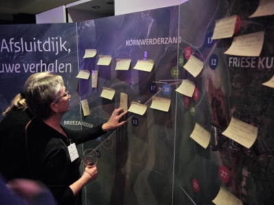 Ondernemer praat mee over de ontwikkelingen bij de Afsluitdijk. Een mevrouw staat voor een bord met de kaart van de Afsluitdijk. Zij plakt gele memokaartjes op het bord.