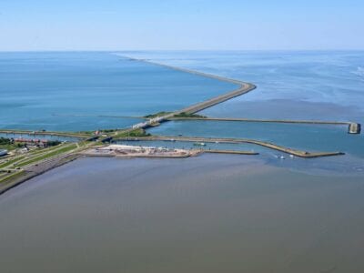 Luchtfoto van het Afsluitdijk Wadden Center, deze ligt links. De weg van de Afsluitdijk loopt van linksonder met een bocht naar linksboven. In het midden zien we de huizen van Kornwerderzand. Daarnaast het kantoor van DNA. Je ziet de weg die over de Afsluitdijk loopt. Aan de beide kanten van de weg zie je water.