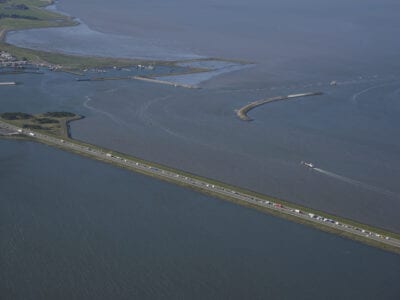 Luchtfoto van Den Oever. We zien links een klein stukje van Den Oever en de haven. Er loopt een weg rechtsonder uit beeld. Deze is omringt door water. Boven in beeld zie je een gebogen landstrook. Dit is de dam: de banaan.