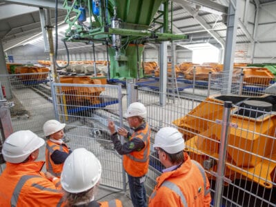 Wij staan binnen in een loods. Daar zien wij de openingshandeling door Avine Fokkens. Zij staat voor een hek met een aantal werkmannen. Op de achtergrond staan veel gele blokken.