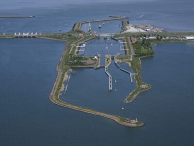Wij zien een luchtfoto van de Schutsluizen bij Kornwerderzand. Van links naar rechts loopt een weg. Verticaal lopen vier landstroken met sluizen en boten. Aan beide kanten van de weg zie je water.