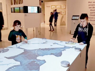 We zien een tafel in het Afsluitdijk Wadden Center. Op de tafel zie je een blauwe weergave van een stuk land. Op de tafel staan ook een paar knoppen en twee kindjes spelen er mee. Op de achtergrond zie je witte wanden met teksten.