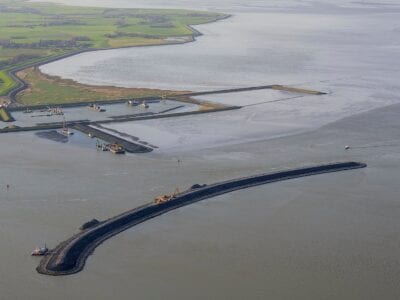 Je ziet de Dam de Banaan in het water. Dit is een gebogen landstrook in het water. Op de achtergrond zie je Den Oever liggen en de haven.