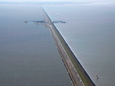 Luchtfoto van de weg op de Afsluitdijk. De weg loopt van beneden naar boven. In de verte zie je een stukje van Breezanddijk. Dit is een stukje land aan de weg.