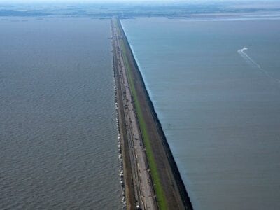 Luchtfoto van de Afsluitdijk. Wij kijken van boven op de weg, vanaf Den Oever. Je ziet de weg met aan beide kanten water. In de verte zie je een stukje van Fryslân.