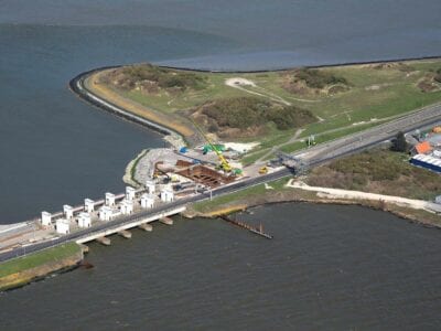 Luchtfoto van de Afsluitdijk. Wij kijken van boven op de Afsluitdijk bij de witte, voormalige sluisgebouwen, Gates Of Light. Je ziet boven en onder een klein stukje van de Waddenzee en het IJsselmeer. Links zie je de witte gebouwen van Gates of Lights. Rechts naast de witte gebouwen, zie je een graslandschap. Onder het landschap zie je een weg lopen. Daaronder zie je een blauw gebouw van Blue Energy.