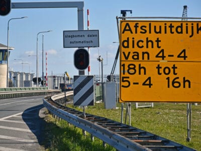 Op de foto zie je het begin van de Afsluitdijk. Aan de weg zie je een verdrijvingsvlak. Aan de zijkant zie je een sluisgebouw en een slagboom. Aan de zijkant zie je een wit bord met de tekst: Slagbomen dalen automatisch. Voorin staat een geel bord over de afsluiting: Afsluitdijk dicht van 4-4 18uur tot 5-4 16uur
