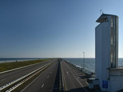 Op de foto zie je de Afsluitdijk. De foto is schuin naar onder gemaakt. Je ziet drie wegdelen en aan de rechterkant het Vlietermonument. Dit monument is een wit uitkijkgebouw. Bovenaan het gebouw zie je een opening waar mensen over de dijk kunnen kijken. Aan de rechterkant van het gebouw zie je een glazenmuur met op de achtergrond een wenteltrap.