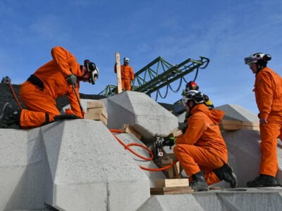 Veiligheidsoefening met Levvelblocs. Je ziet drie personen in oranje pakken knielen bij de blokken. Deze blokken zijn in een hoekige vorm gemaakt. Zij plaatsen een hefboom-systeem tussen de blokken. En liften de blokken zo omhoog. Boven de blokken zie je de lucht en een hijskraan.