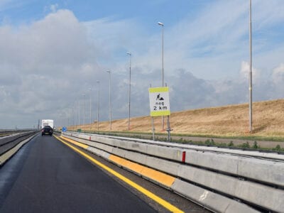 Je ziet een weg die versmald is met behulp van een metalen railing en een gele doorgetrokken streep. Aan de rechterkant van de weg staat een bord met de tekst nog 2 kilometer.
