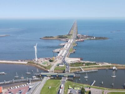 Op de foto vanuit de lucht zie je de dijk van bovenaf die van onder naar boven loopt. In het midden van de foto zie je de heftorens van een spuicomplex. Het verkeer rijdt over de rechter rijbaan. De linker rijbaan is afgesloten.