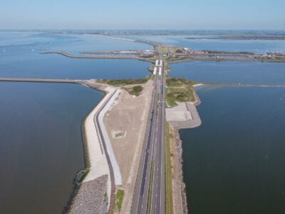 Je ziet een luchtfoto van de Afsluitdijk. Der weg loopt recht naar boven met aan het einde een bocht naar links. De Afsluitdijk wordt omringt door water.