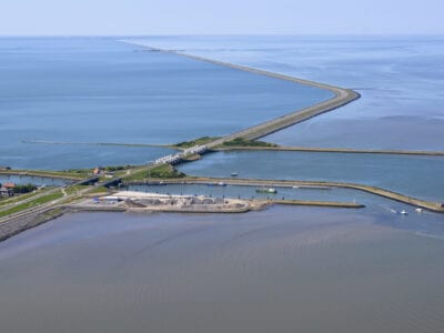 Op de foto zie je de nieuwe sluis van Kornwerderzand. De foto is van bovengenomen. Door het beeld loop de weg van de Afsluitdijk. Aan beide kanten van de Afsluitdijk zie je groen en water.