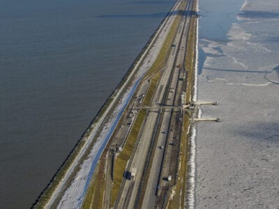 Luchtfoto van de Afsluitdijk in de winter. We kijken uit over het monument, de dijk en het water.