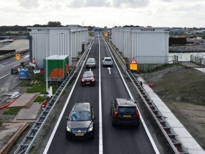 We zien de weg van de Afsluitdijk. Er rijden meerdere auto's overheen.