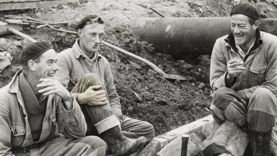 zwart wit foto uit de jaren 1920. Drie werklieden van de Afsluitdijk zitten op de basaltblokken en lachen met elkaar. Op de achtergrond zie je de bouwplaats.