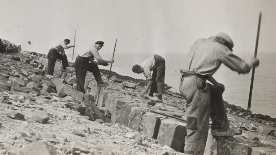 Zwart wit foto van vier mannen die aan het werk zijn met de aanleg van een dijk met grote basaltblokken.