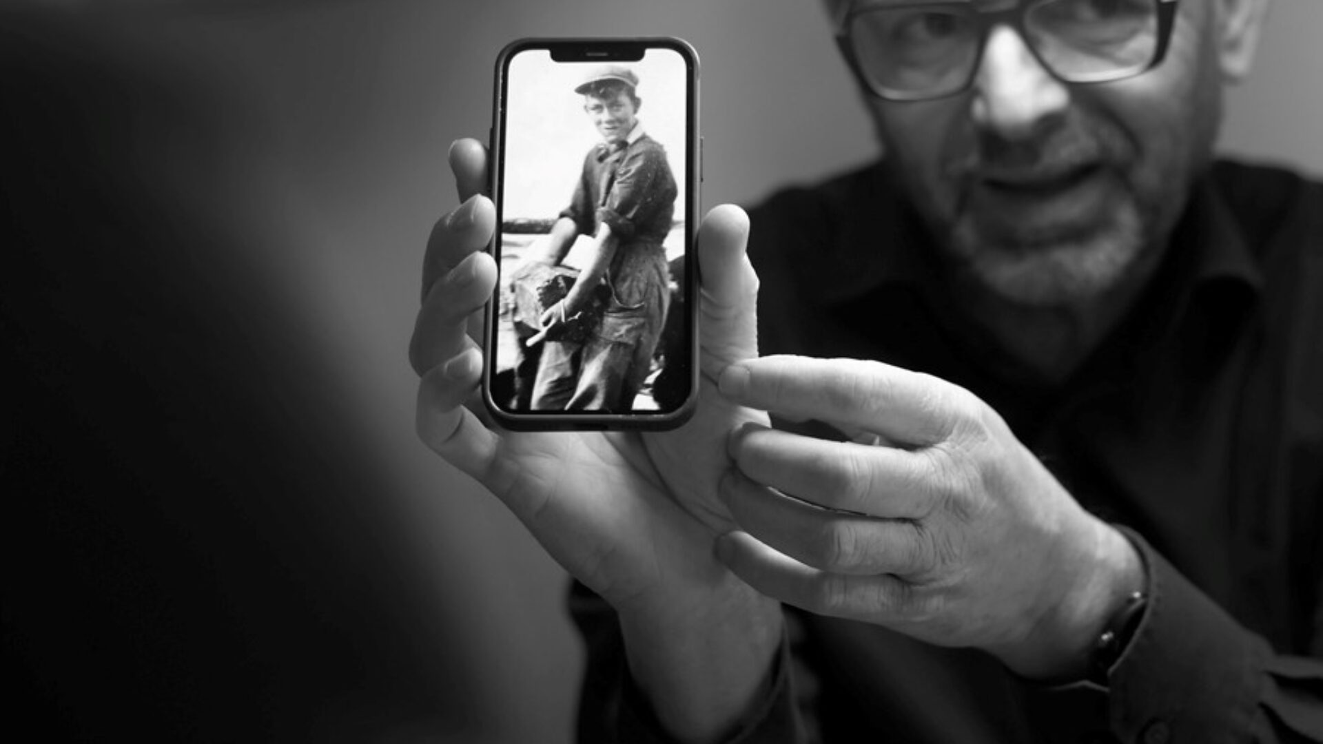 Zwart wit foto van iemand die naar de camera kijkt en een foto toont op zijn mobiel. Daarop is een oude foto te zien van een voorvader aan het werk aan de Afsluitdijk.