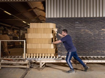 Een kar met stapels lichtgele tegels wordt in een loods weggereden door een medewerker.