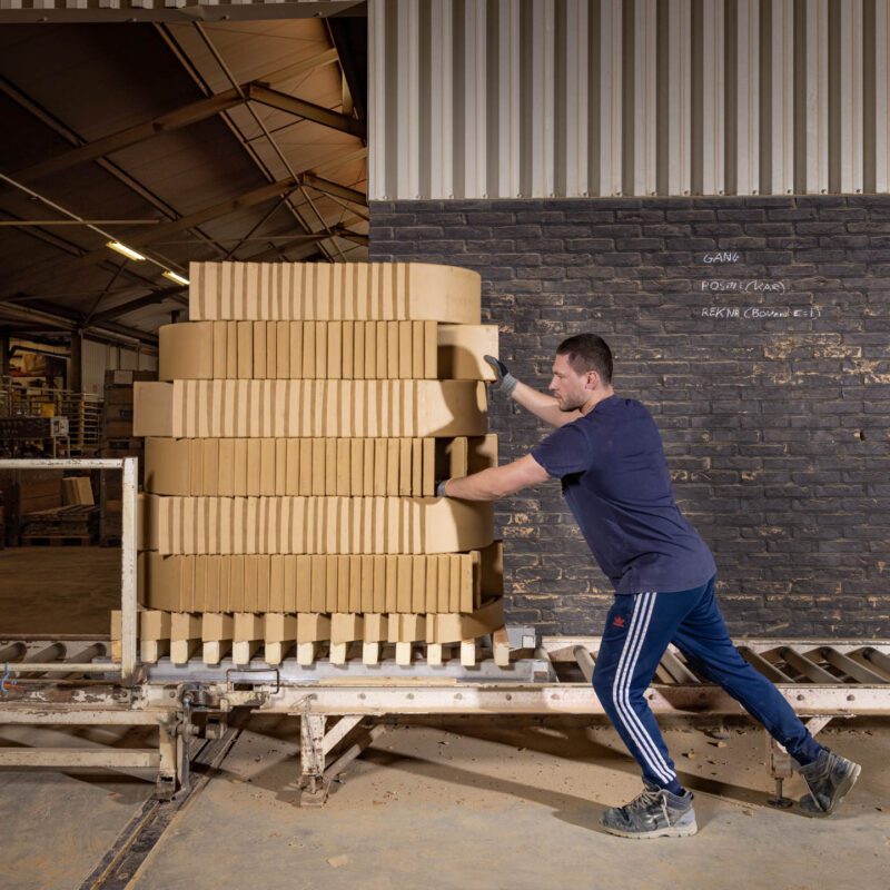 Een kar met stapels lichtgele tegels wordt in een loods weggereden door een medewerker.