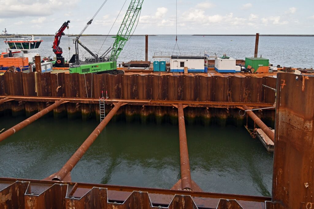 Stalen wanden bij een bouwproject. De wanden worden stabiel gehouden door grote stalen buizen. Tussen de wanden staat water. 