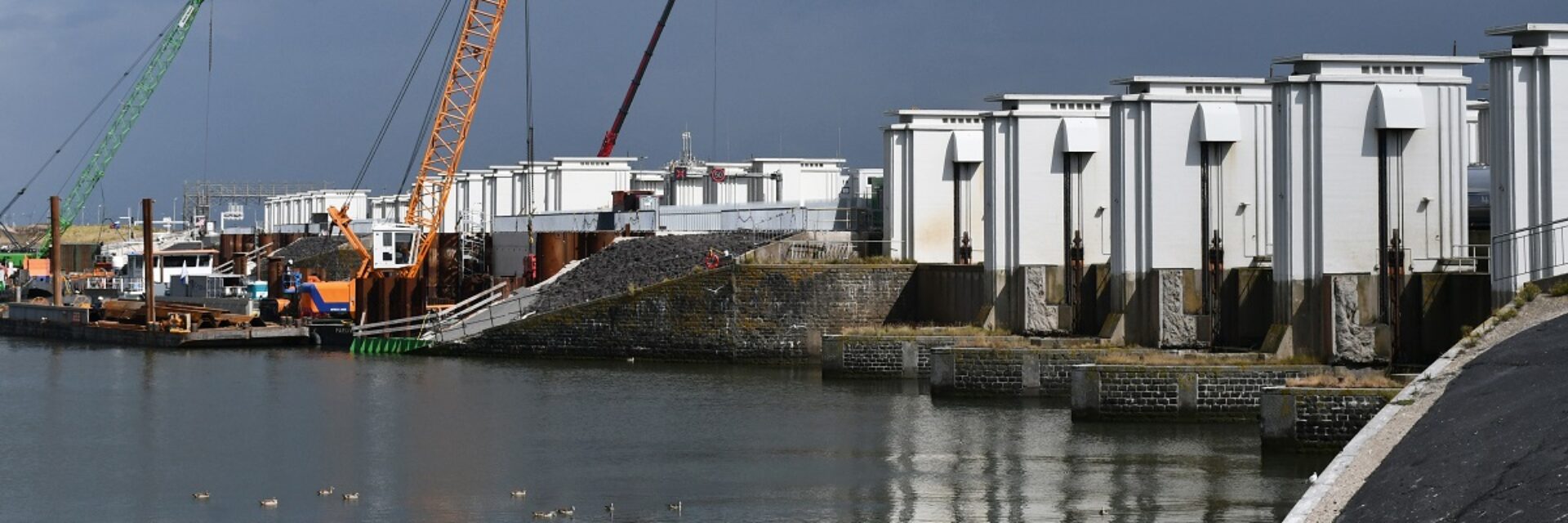 Witte torentjes rechts op een rij op de Afsluitdijk met links ervoor water en aan de horizon een aantal kraanschepen.