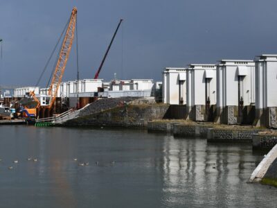 Witte torentjes rechts op een rij op de Afsluitdijk met links ervoor water en aan de horizon een aantal kraanschepen.