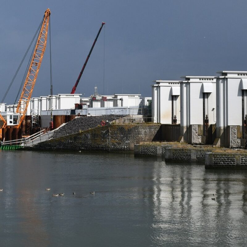 Witte torentjes rechts op een rij op de Afsluitdijk met links ervoor water en aan de horizon een aantal kraanschepen.